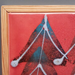 May include: A framed ceramic tile with a vibrant red background. The tile features a geometric design in teal, black, and white. The frame is made of light-coloured wood. This artwork is suitable for wall decor.