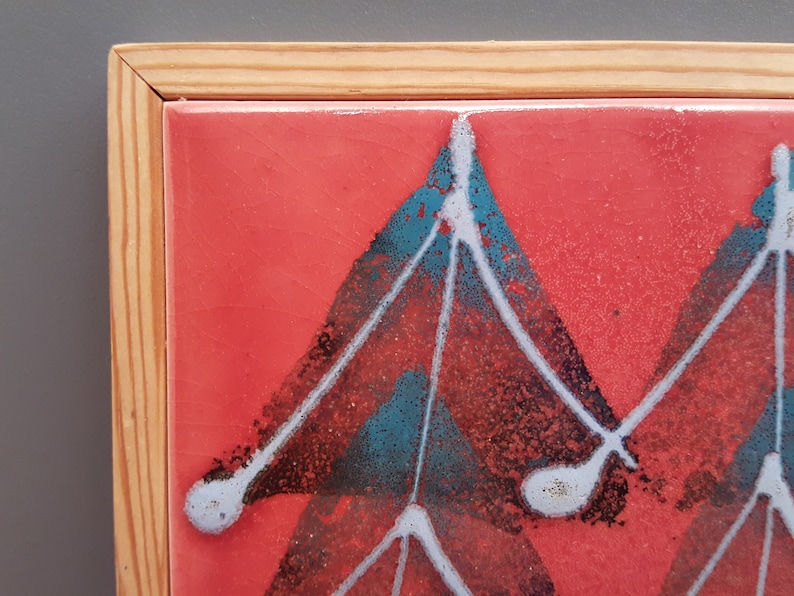 May include: A framed ceramic tile with a vibrant red background. The tile features a geometric design in teal, black, and white. The frame is made of light-coloured wood. This artwork is suitable for wall decor.
