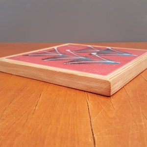 May include: Square coaster with a red surface and a geometric design of white and teal shapes. The coaster has a natural wood edge and is set on a wooden table.