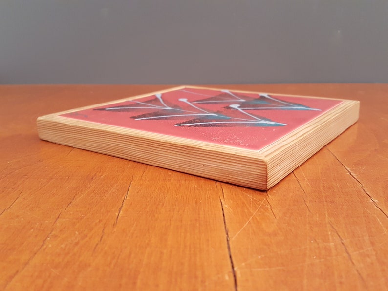 May include: Square coaster with a red surface and a geometric design of white and teal shapes. The coaster has a natural wood edge and is set on a wooden table.