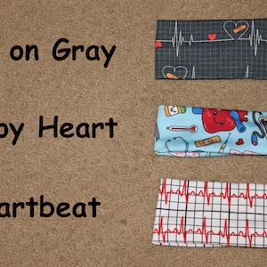 May include: Three headbands with different medical-themed prints. The top headband has a black and white EKG pattern with red hearts. The middle headband has a light blue background with a red heart, a stethoscope, a thermometer, and other medical symbols. The bottom headband has a white background with a red EKG pattern.