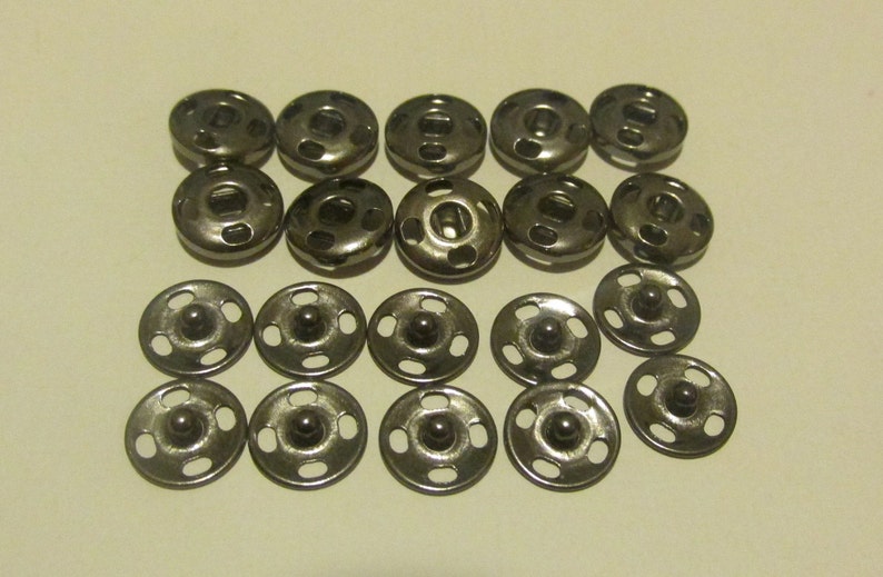 May include: A set of 18 silver metal snap fasteners. The fasteners are round and have a raised center. They are arranged in two rows of nine.
