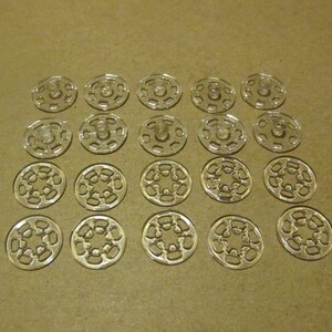 May include: A collection of 18 clear plastic snap fasteners, arranged in a grid pattern on a brown surface. Each fastener has a central hole and a series of smaller holes around the edge.