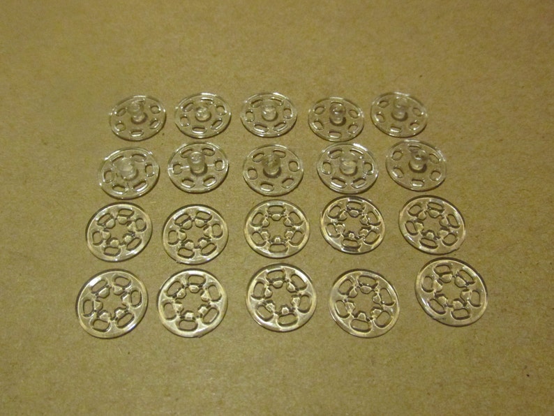 May include: A collection of 18 clear plastic snap fasteners, arranged in a grid pattern on a brown surface. Each fastener has a central hole and a series of smaller holes around the edge.