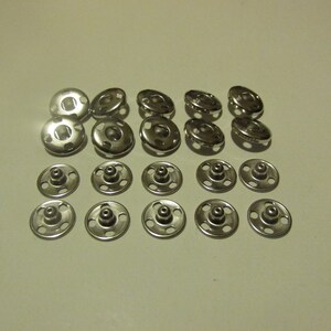 May include: A set of 20 silver metal snap fasteners. The fasteners are arranged in two rows, with 10 fasteners in each row.