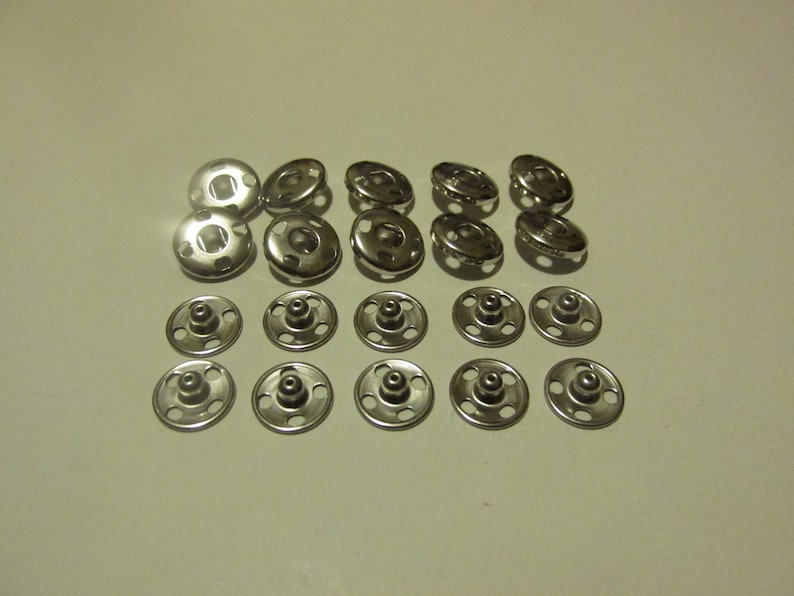 May include: A set of 20 silver metal snap fasteners. The fasteners are arranged in two rows, with 10 fasteners in each row.