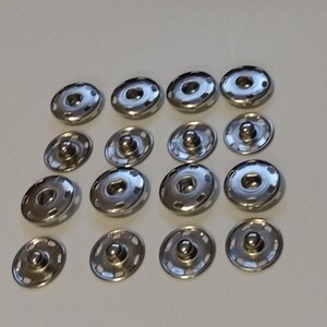 May include: A set of 18 silver metal snap fasteners. The fasteners are round and have a raised center. They are arranged in three rows of six.