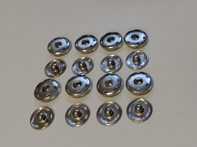 May include: A set of 18 silver metal snap fasteners. The fasteners are round and have a raised center. They are arranged in three rows of six.