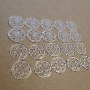 May include: A set of 18 clear plastic snap fasteners. The fasteners are arranged in two rows of nine. The fasteners are designed to be used for attaching fabric or other materials together.
