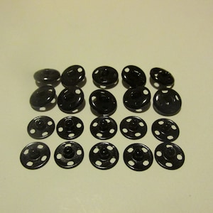 May include: A set of 20 black metal snap fasteners. The fasteners are round and have four holes around the edge. They are commonly used for clothing and other fabric projects.