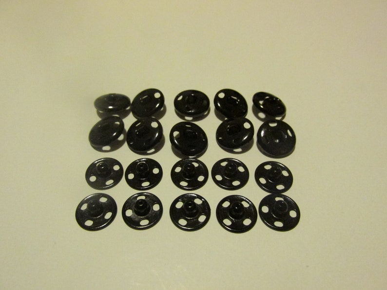 May include: A set of 20 black metal snap fasteners. The fasteners are round and have four holes around the edge. They are commonly used for clothing and other fabric projects.