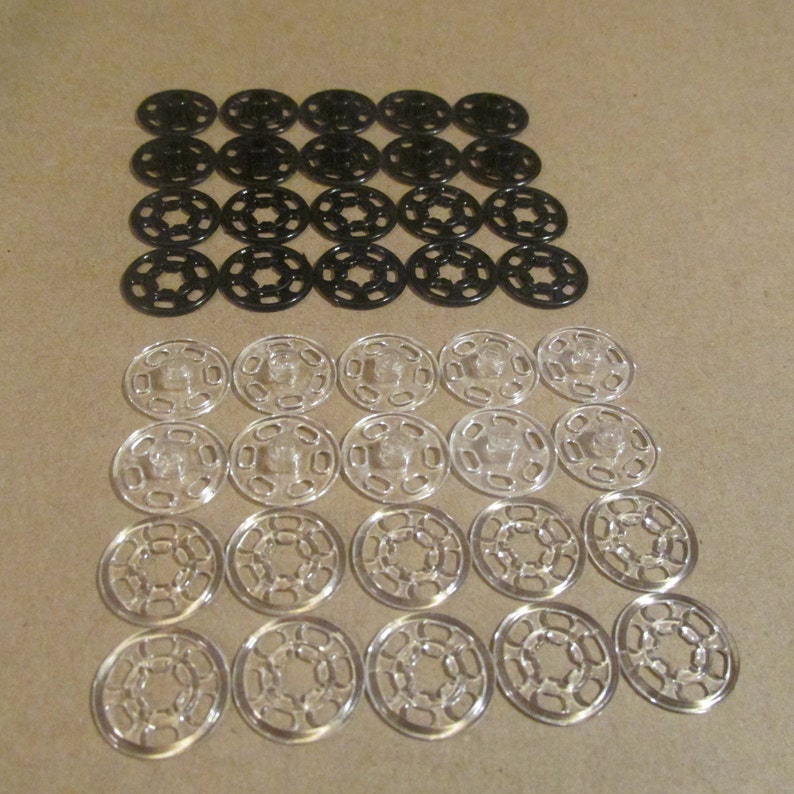 May include: A set of 24 snap fasteners, 12 black and 12 clear. The fasteners are arranged in two rows of 12, with the black fasteners on top and the clear fasteners on the bottom.