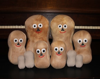 May include: A group of seven plush peanut toys, all with smiling faces and large eyes. The peanuts are arranged in a pyramid shape, with the largest peanut at the top and the smallest peanuts at the bottom.