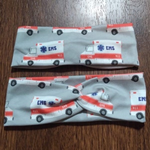 May include: Two gray fabric headbands with a repeating pattern of white and red ambulance illustrations. The ambulances have the text "EMS 911" on them.