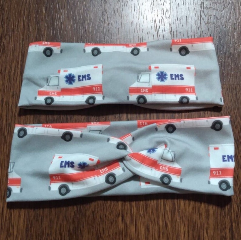 May include: Two gray fabric headbands with a repeating pattern of white and red ambulance illustrations. The ambulances have the text "EMS 911" on them.