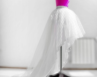 Puede incluir: Una falda de tul blanca con un dobladillo alto-bajo, expuesta en un maniquí rosa. La falda es fluida y tiene una textura suave y delicada.