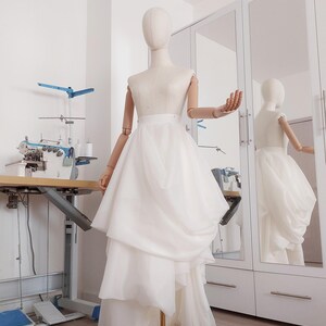 Op de afbeelding: Een witte trouwjurk op een mannequin. De jurk heeft een gelaagde rok en een getailleerd lijfje. De mannequin staat in een studio met een naaimachine en een spiegel.