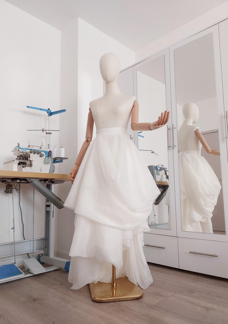 Op de afbeelding: Een witte trouwjurk op een mannequin. De jurk heeft een gelaagde rok en een getailleerd lijfje. De mannequin staat in een studio met een naaimachine en een spiegel.