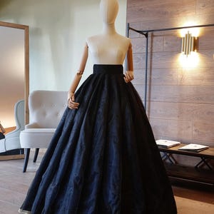 Op de afbeelding: Een zwarte, vloerlange rok met een brede tailleband en een volle, vloeiende silhouet. De rok wordt getoond op een mannequin met een crèmekleurig lijfje. De zoom is afgezet met een goudkleurige rand. De rok is gemaakt van een transparante stof.