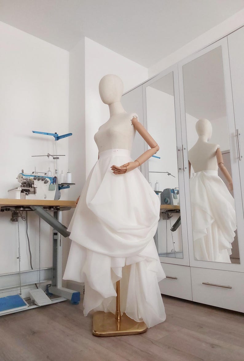Op de afbeelding: Een witte trouwjurk op een paspop. De jurk heeft een getailleerd lijfje en een gelaagde rok met een uniek gedrapeerd ontwerp. De paspop staat in een lichte kamer met een naaimachine en een spiegel.