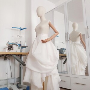 Op de afbeelding: Een witte trouwjurk op een mannequin in een studio. De jurk heeft een getailleerd lijfje en een gelaagde, asymmetrische rok. De mannequin staat voor een spiegel. Een naaimachine en andere gereedschappen zijn zichtbaar op de achtergrond.