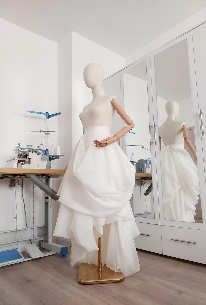 Op de afbeelding: Een witte trouwjurk op een mannequin in een studio. De jurk heeft een getailleerd lijfje en een gelaagde, asymmetrische rok. De mannequin staat voor een spiegel. Een naaimachine en andere gereedschappen zijn zichtbaar op de achtergrond.