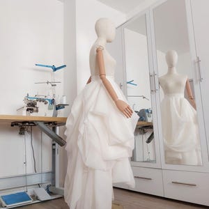 Op de afbeelding: Een witte trouwjurk op een mannequin, met een gelaagde rok en een nauwsluitend lijfje. De jurk wordt getoond in een lichte kamer met een naaimachine en een grote spiegel die de jurk weerspiegelt. De mannequin heeft beweegbare armen.