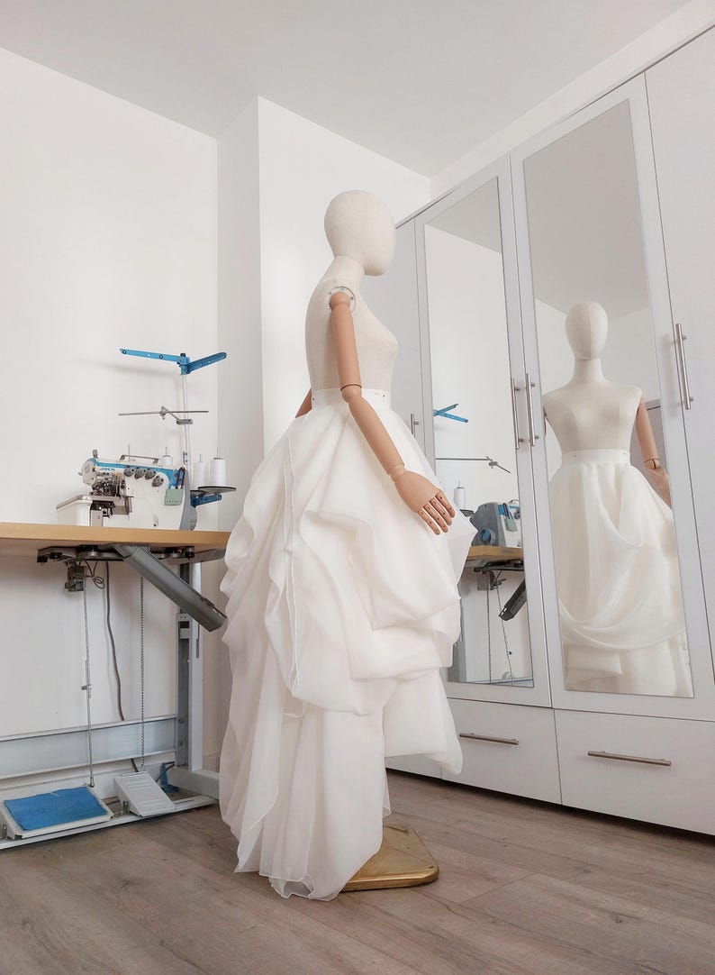 Op de afbeelding: Een witte trouwjurk op een mannequin, met een gelaagde rok en een nauwsluitend lijfje. De jurk wordt getoond in een lichte kamer met een naaimachine en een grote spiegel die de jurk weerspiegelt. De mannequin heeft beweegbare armen.