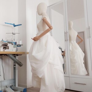 Op de afbeelding: Een witte trouwjurk op een mannequin, met een gelaagde rok en een nauwsluitend lijfje. De jurk wordt getoond in een lichte kamer met een naaimachine en een spiegel die de jurk weerspiegelt. De mannequin heeft houten armen.
