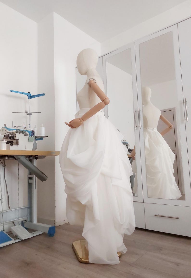 Op de afbeelding: Een witte trouwjurk op een mannequin, met een gelaagde rok en een nauwsluitend lijfje. De jurk wordt getoond in een lichte kamer met een naaimachine en een spiegel die de jurk weerspiegelt. De mannequin heeft houten armen.