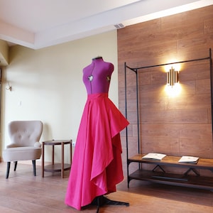 May include: A magenta dress form with a vibrant pink skirt. The form is set in a room with wood flooring and a wooden accent wall. A small chair and side table are in the background.