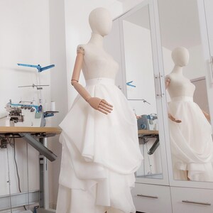 Op de afbeelding: Een witte trouwjurk op een mannequin. De jurk heeft een gelaagde rok en een getailleerd lijfje. De mannequin staat in een lichte kamer met een naaimachine en een spiegel.
