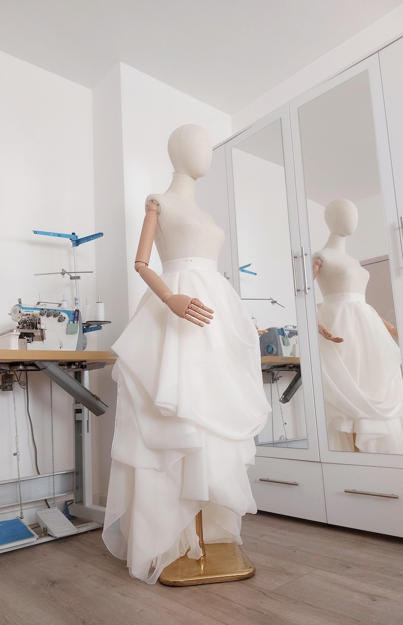Op de afbeelding: Een witte trouwjurk op een mannequin. De jurk heeft een gelaagde rok en een getailleerd lijfje. De mannequin staat in een lichte kamer met een naaimachine en een spiegel.