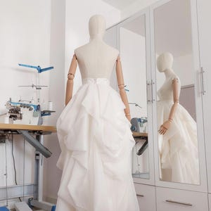 Op de afbeelding: Een witte trouwjurk op een mannequin in een bruidswinkel. De jurk heeft een gelaagde rok en een getailleerd lijfje. Een naaimachine en een spiegel zijn zichtbaar op de achtergrond, wat duidt op een ontwerp op maat.
