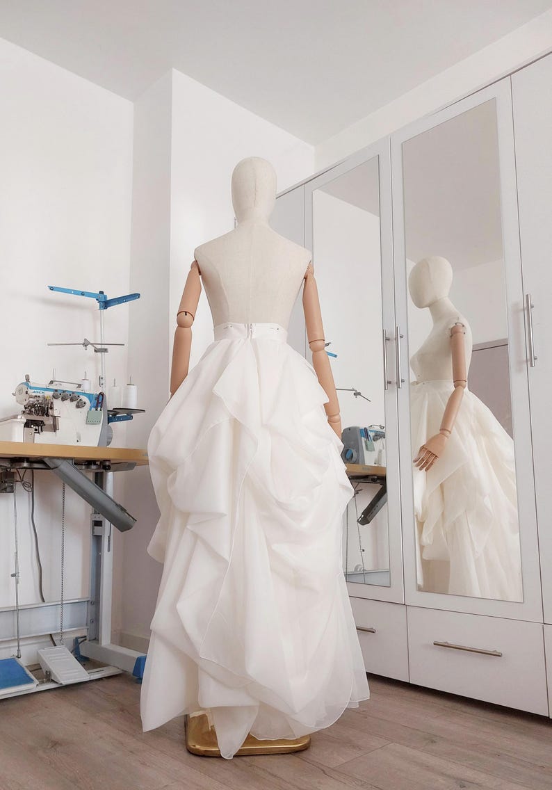 Op de afbeelding: Een witte trouwjurk op een mannequin in een bruidswinkel. De jurk heeft een gelaagde rok en een getailleerd lijfje. Een naaimachine en een spiegel zijn zichtbaar op de achtergrond, wat duidt op een ontwerp op maat.