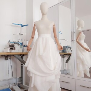 Op de afbeelding: Een witte trouwjurk op een paspop. De jurk heeft een gelaagde rok en een getailleerd lijfje. De paspop heeft houten armen en een neutrale kop. De jurk wordt getoond in een lichte kamer met een naaimachine en een spiegel.