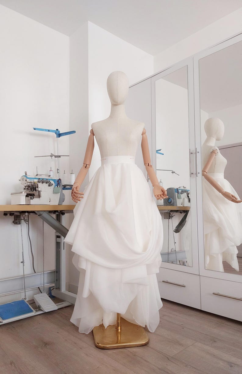 Op de afbeelding: Een witte trouwjurk op een paspop. De jurk heeft een gelaagde rok en een getailleerd lijfje. De paspop heeft houten armen en een neutrale kop. De jurk wordt getoond in een lichte kamer met een naaimachine en een spiegel.