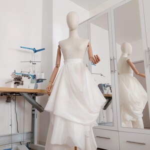 Op de afbeelding: Een witte gelaagde rok op een mannequin. De rok heeft een high-low zoomlijn en wordt tentoongesteld in een goed verlichte kamer met een naaimachine en een spiegel. De mannequin is van hout en heeft beweegbare armen.