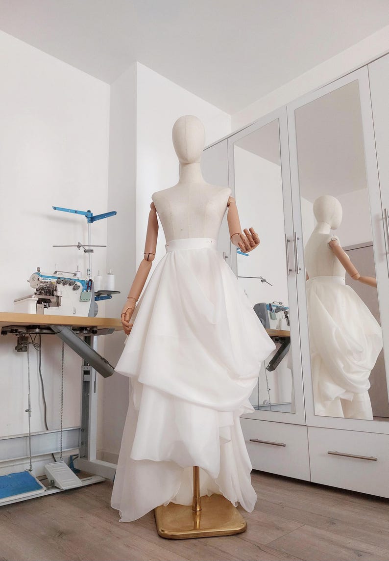 Op de afbeelding: Een witte gelaagde rok op een mannequin. De rok heeft een high-low zoomlijn en wordt tentoongesteld in een goed verlichte kamer met een naaimachine en een spiegel. De mannequin is van hout en heeft beweegbare armen.