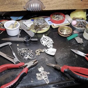 May include: A cluttered workbench with jewelry-making tools and components. The image shows various pliers, metal pendants, small containers with findings, and a drawer pull. The tools are arranged on a dark surface, suggesting a workspace for crafting.