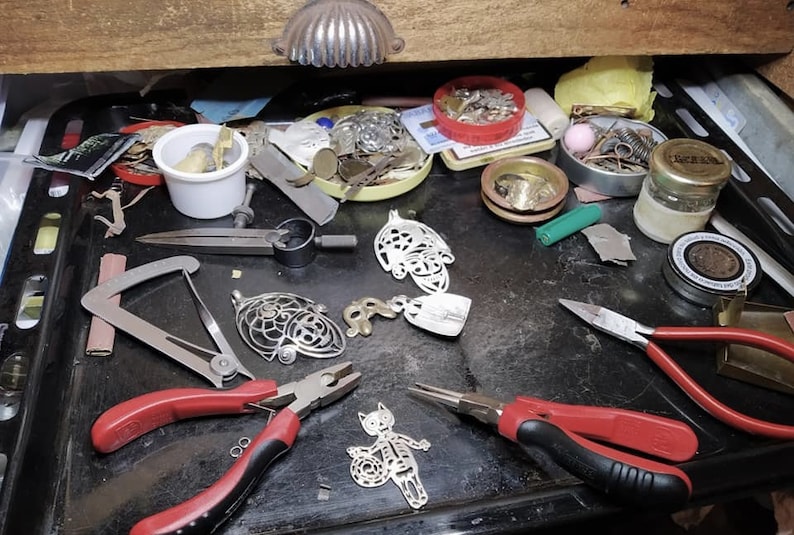 May include: A cluttered workbench with jewelry-making tools and components. The image shows various pliers, metal pendants, small containers with findings, and a drawer pull. The tools are arranged on a dark surface, suggesting a workspace for crafting.