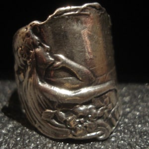 May include: A silver-toned ring with an embossed design featuring a classical female figure and floral elements. The ring has a wide band and a detailed, textured surface. The design wraps around the ring, showcasing the figure's profile and surrounding floral details.