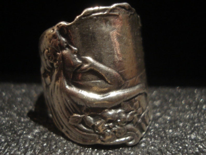 May include: A silver-toned ring with an embossed design featuring a classical female figure and floral elements. The ring has a wide band and a detailed, textured surface. The design wraps around the ring, showcasing the figure's profile and surrounding floral details.