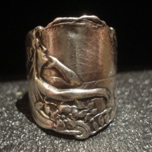 May include: A silver ring with an embossed design featuring a reclining figure, flowers, and foliage. The ring has a cylindrical shape and a detailed, antique appearance. The ring is likely made of sterling silver or a similar metal.