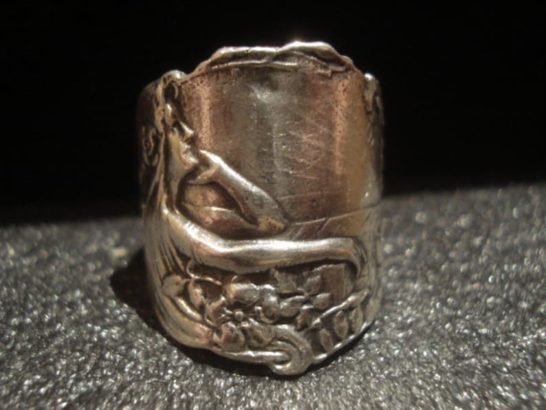 May include: A silver ring with an embossed design featuring a reclining figure, flowers, and foliage. The ring has a cylindrical shape and a detailed, antique appearance. The ring is likely made of sterling silver or a similar metal.