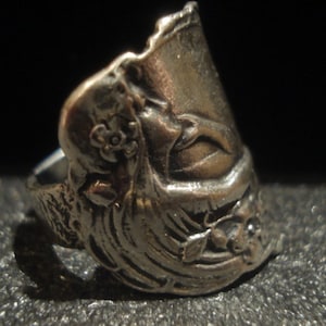 May include: A silver-toned ring with an ornate design. The ring features floral and face details, with a textured surface. The ring is set against a dark background, highlighting its intricate craftsmanship.