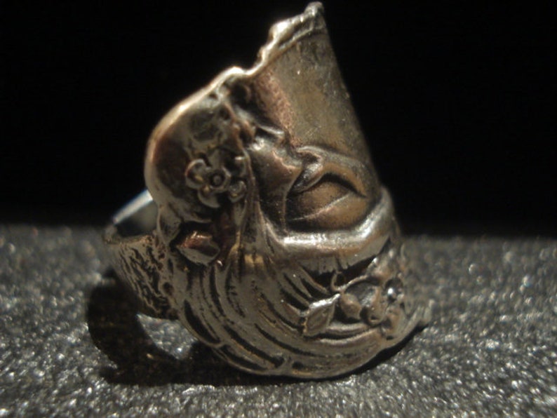 May include: A silver-toned ring with an ornate design. The ring features floral and face details, with a textured surface. The ring is set against a dark background, highlighting its intricate craftsmanship.