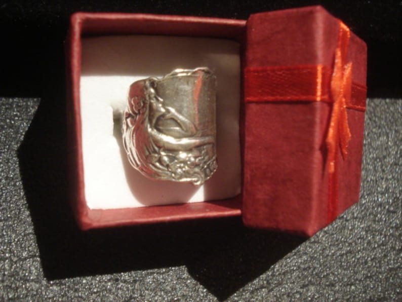 May include: A silver ring with an ornate design, presented in a small red gift box with a red ribbon. The ring features a detailed, possibly antique, design. The box is open, revealing the ring on a white interior.