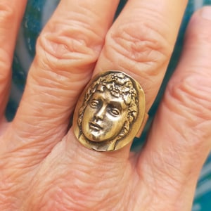 Puede incluir: Un anillo de tono dorado con un diseño ovalado detallado que presenta una cara con una corona de laurel. El anillo se lleva en un dedo.