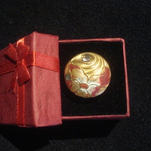 Puede incluir: Un adorno redondo dorado y rojo con un diseño floral y un solo strass en el centro. El adorno está en una caja de regalo roja con una cinta roja.